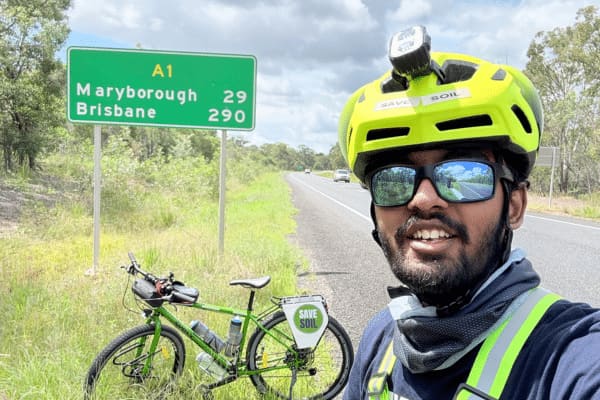 Sahil Jha taking a selfie beside a road sign for Maryborough and Brisbane, his bicycle parked behind him