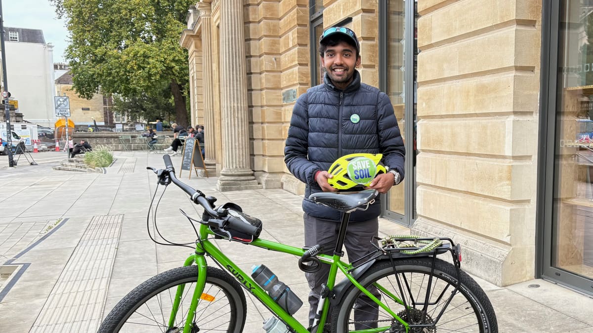 Sahil Jha smiling beside his green touring bicycle, holding a helmet with a Save Soil sticker, on a stone-paved street in the United Kingdom